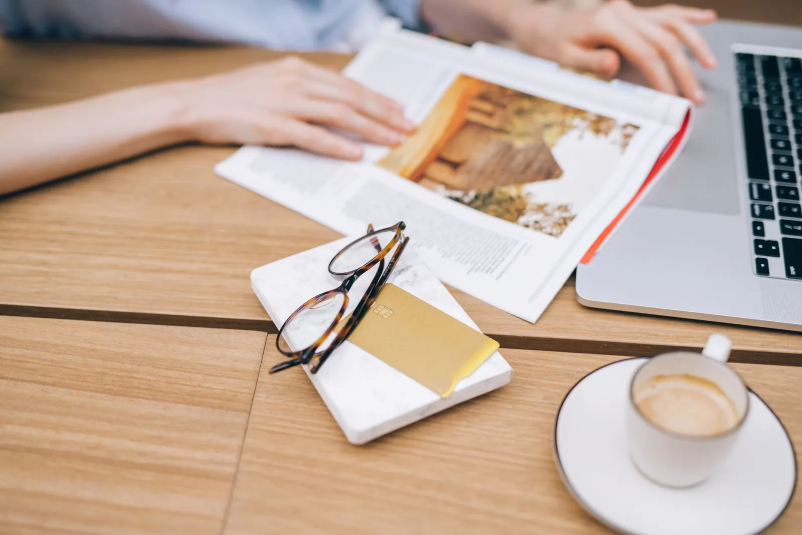 A warm workspace featuring a laptop, coffee cup, magazine, and eyeglasses on a wooden table.