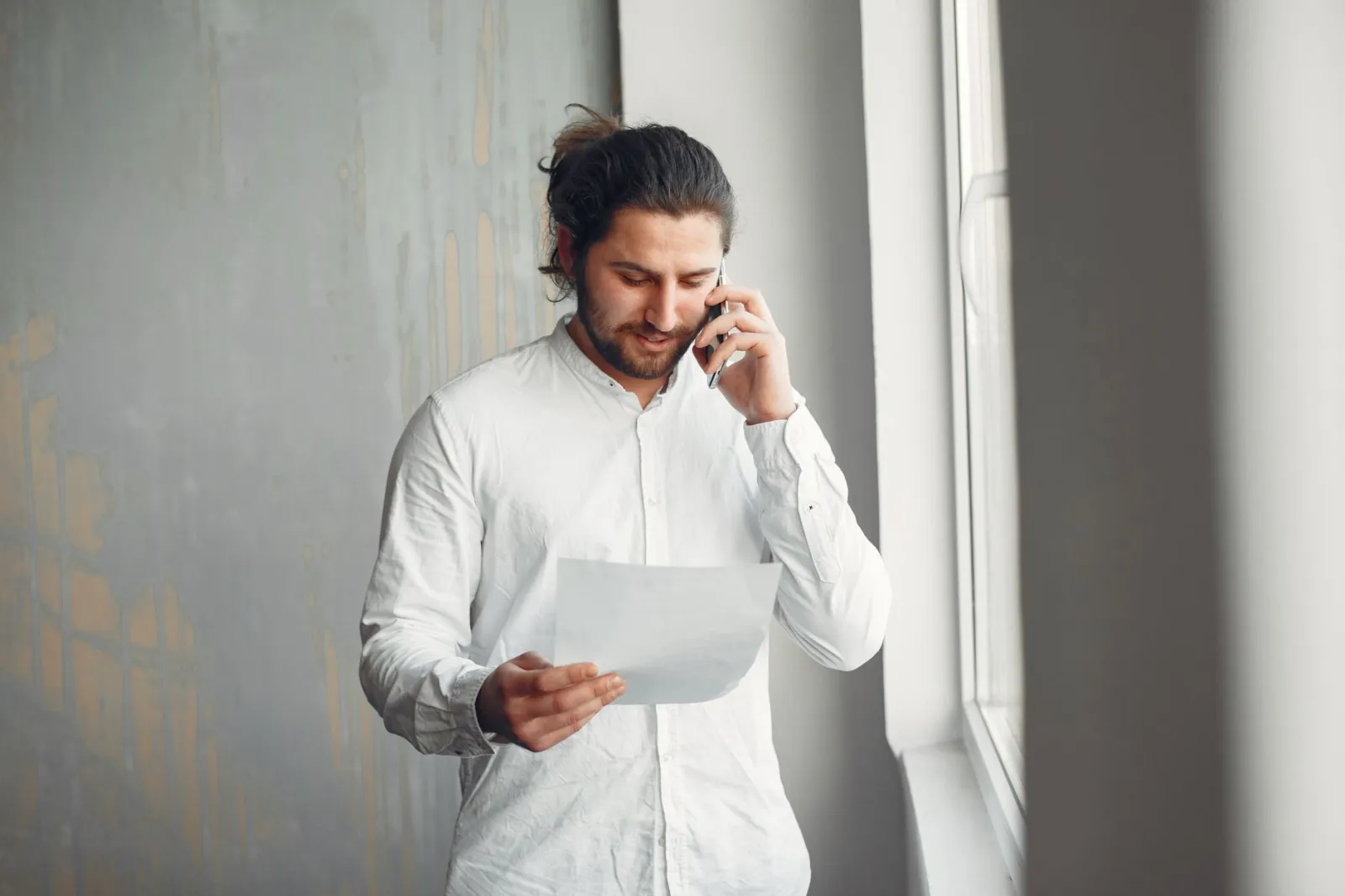 A young man in casual attire talks on the phone while holding a document indoors.
